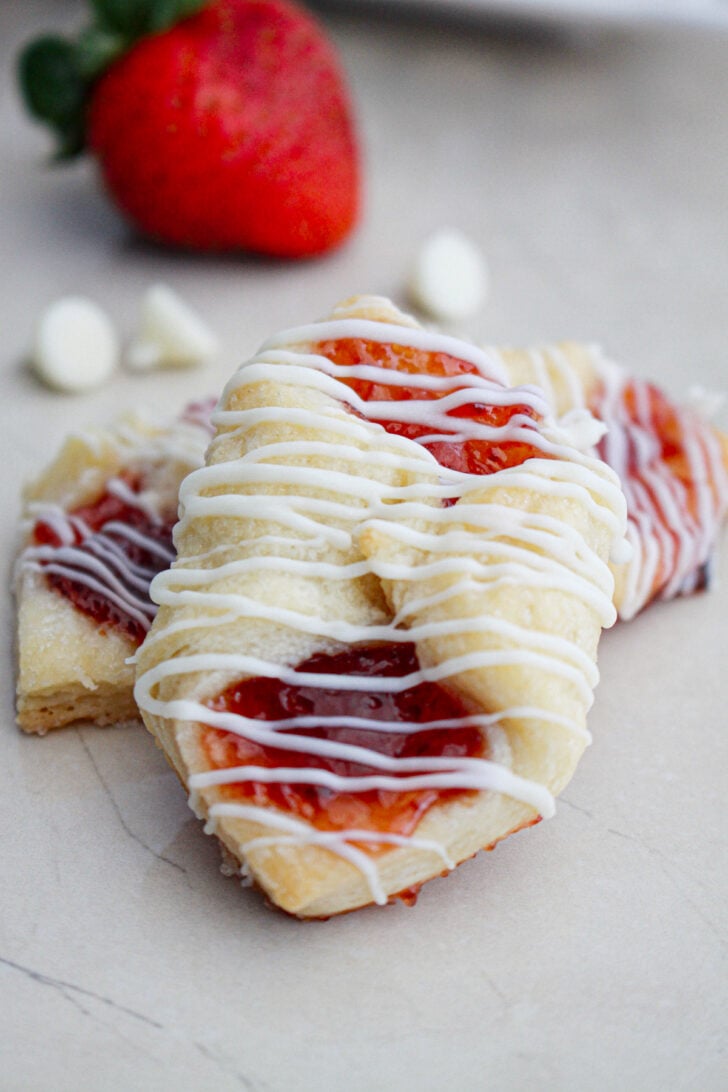 Two pastries with jam filling and white icing drizzle are stacked on a light surface. A fresh strawberry and a couple of white chocolate chips are in the background.