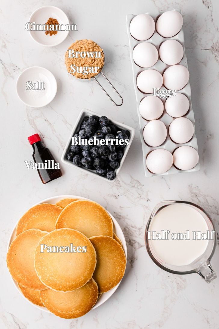 Top view of ingredients for pancakes laid out on a marble surface: a stack of pancakes, blueberries, a dozen eggs, brown sugar, cinnamon, salt, vanilla extract, and a measuring cup of half and half.