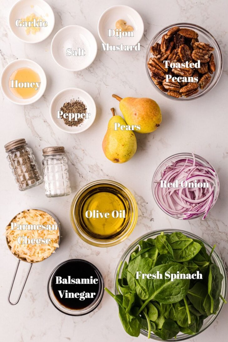 Ingredients for a salad arranged on a white surface: garlic, salt, Dijon mustard, honey, pepper, toasted pecans, pears, red onion, fresh spinach, balsamic vinegar, olive oil, sliced almonds, and grated Parmesan cheese.