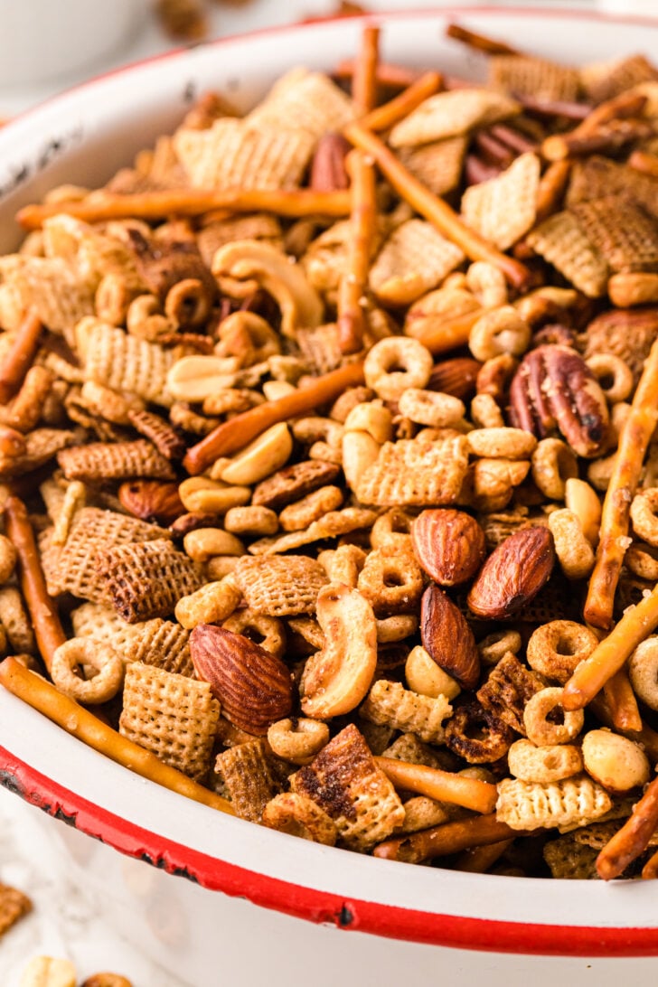 A close-up of a bowl filled with a mix of pretzel sticks, various nuts like almonds and cashews, and assorted cereal pieces. The snack mix is presented in a white bowl with a red rim.