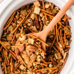 cp party mix 6 in white bowl with wooden spoon showing chex cereal, pretzels, nuts, and almonds mixed together