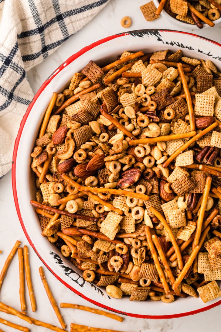 A large bowl filled with assorted snacks, including pretzel sticks, cereal squares, nuts, and cheerios, is placed on a white surface. A checked cloth napkin is beside it, and a few pieces are scattered nearby.