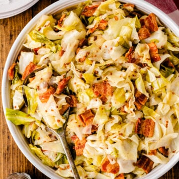 Fried cabbage with crispy bacon pieces served in a white bowl with a spoon, topped with red pepper flakes on wooden surface