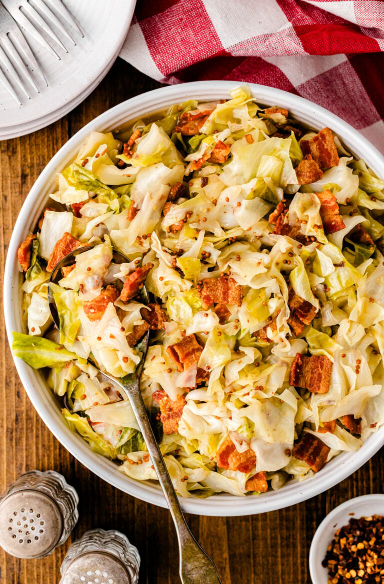 Fried cabbage with crispy bacon pieces served in a white bowl with a spoon, topped with red pepper flakes on wooden surface
