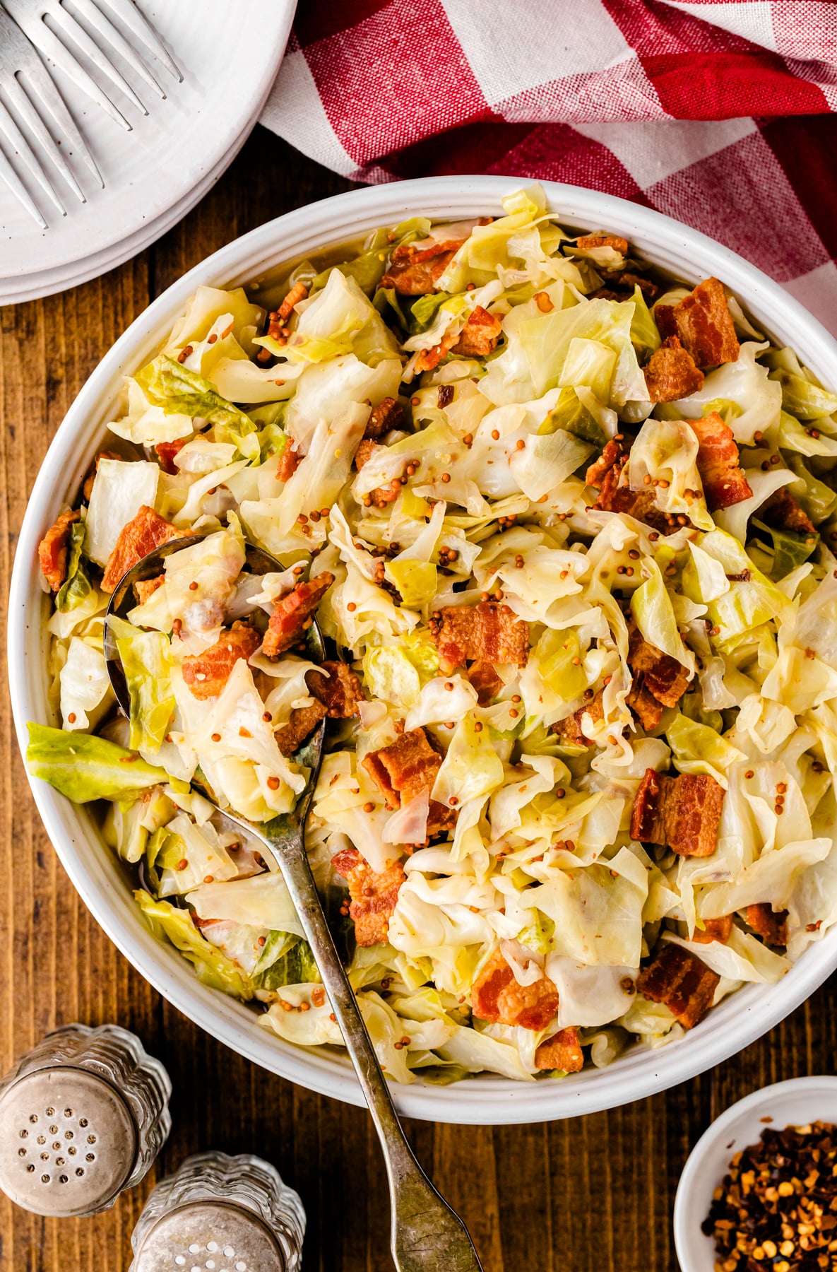 Fried cabbage with crispy bacon pieces served in a white bowl with a spoon, topped with red pepper flakes on wooden surface