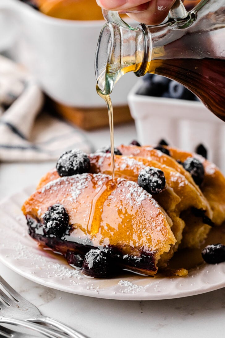 Pancakes topped with blueberries and powdered sugar are on a white plate. Syrup is being poured over them from a glass container. A fork is placed in the foreground, while a bowl of blueberries and a baking dish are blurred in the background.