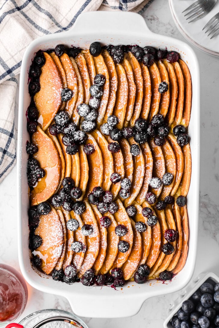 A baked dish of neatly arranged sliced pancakes sprinkled with powdered sugar and topped with blueberries. A checkered cloth and utensils are seen beside the dish on a marble surface.