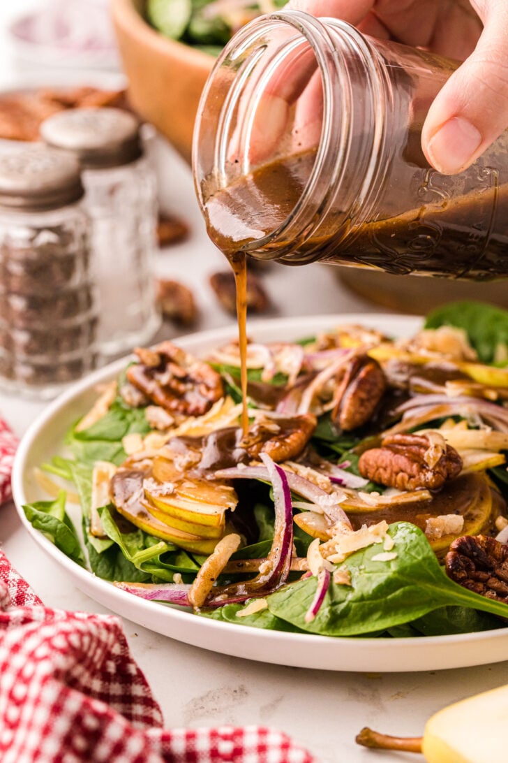 A hand pours vinaigrette dressing from a jar onto a fresh salad with spinach, apple slices, red onion, pecans, and cheese. Salt and pepper shakers, a red-checkered cloth, and a wooden bowl are in the background.