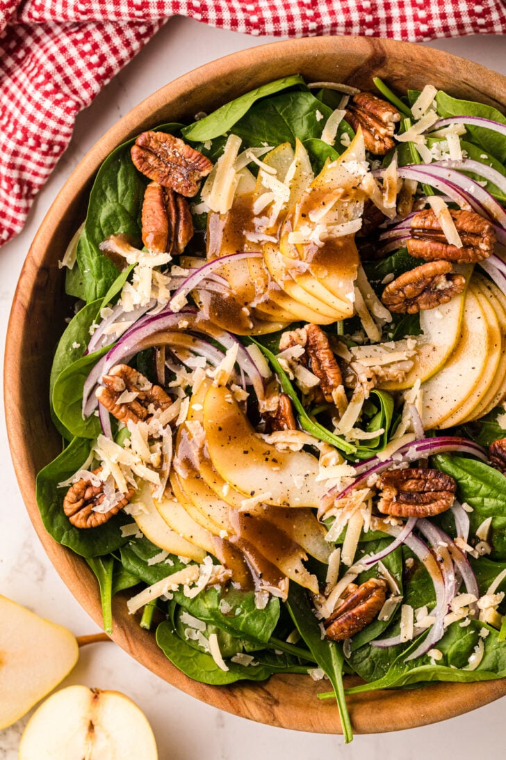 Spinach pear salad 3 in wooden bowl with candied pecans, shredded cheese, red onion, and black pepper, overhead view