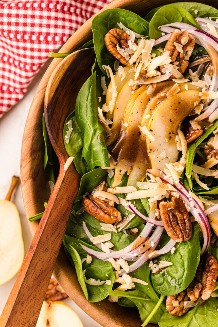 A wooden bowl of fresh spinach salad with sliced pears, pecans, red onions, and grated cheese, topped with a drizzle of balsamic dressing. A wooden spoon and a red-checkered napkin are visible nearby.