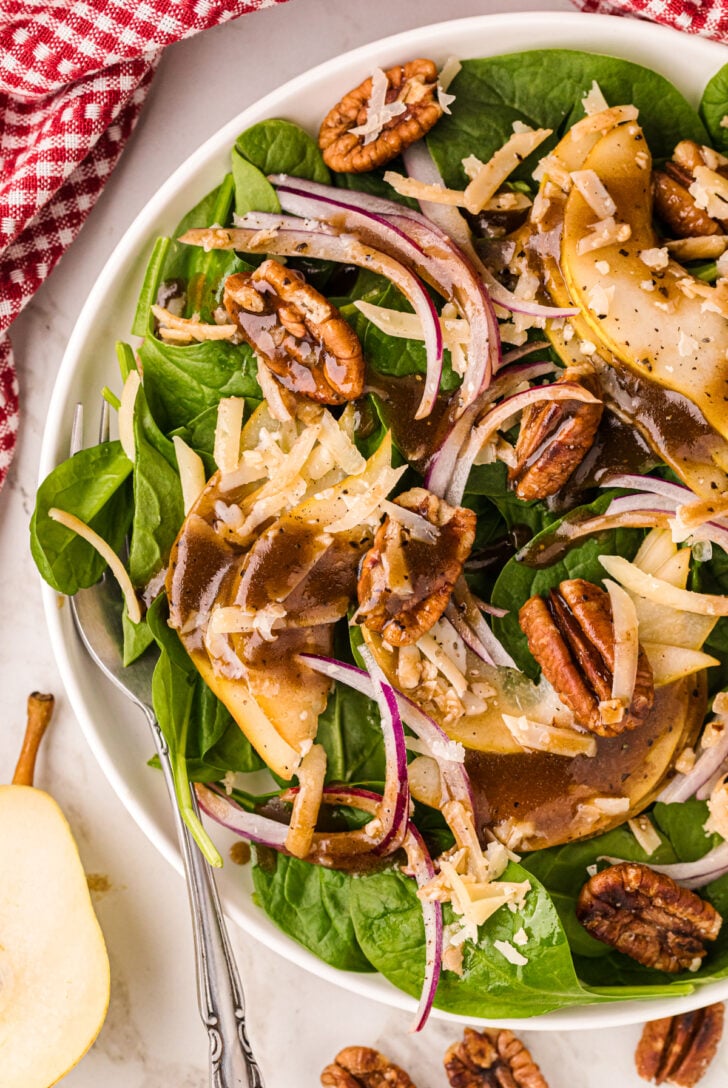 A fresh salad with spinach, red onion slices, pear slices, pecans, and shredded cheese, drizzled with a dark vinaigrette. A red and white checkered cloth is partially visible beside the plate. A pear slice and pecans are on the table.