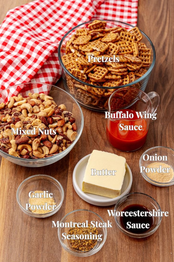 Various snack mix ingredients on a wooden surface include pretzels, mixed nuts, buffalo wing sauce, onion powder, garlic powder, Montreal steak seasoning, Worcestershire sauce, and a block of butter, with a red checkered cloth nearby.