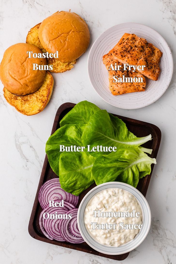 Image of ingredients for a salmon burger. Includes toasted buns, air fryer salmon, butter lettuce, red onions, and a bowl of homemade tartar sauce, all placed on a white surface. Each item is labeled.