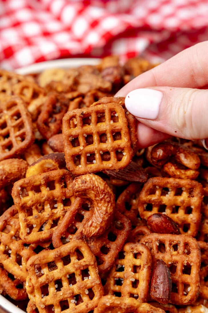 A hand holding a square pretzel above a bowl filled with a snack mix, including square pretzels, almonds, and cashews. A red and white checkered cloth is in the background.