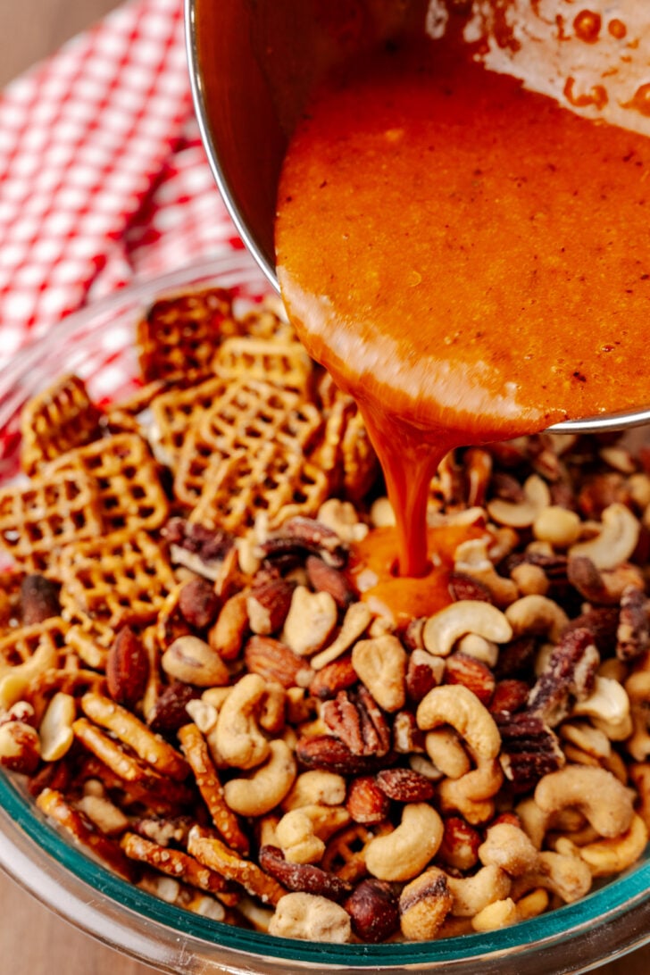 A bowl filled with a mix of nuts and pretzels is being topped with a spicy orange sauce poured from a saucepan. A red and white checkered cloth is in the background.