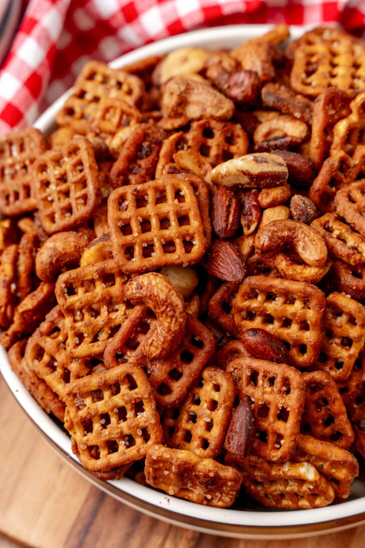 A bowl filled with pretzel crisps and mixed nuts, including almonds and cashews, is placed on a wooden surface. A red and white checkered cloth is partially visible in the background.
