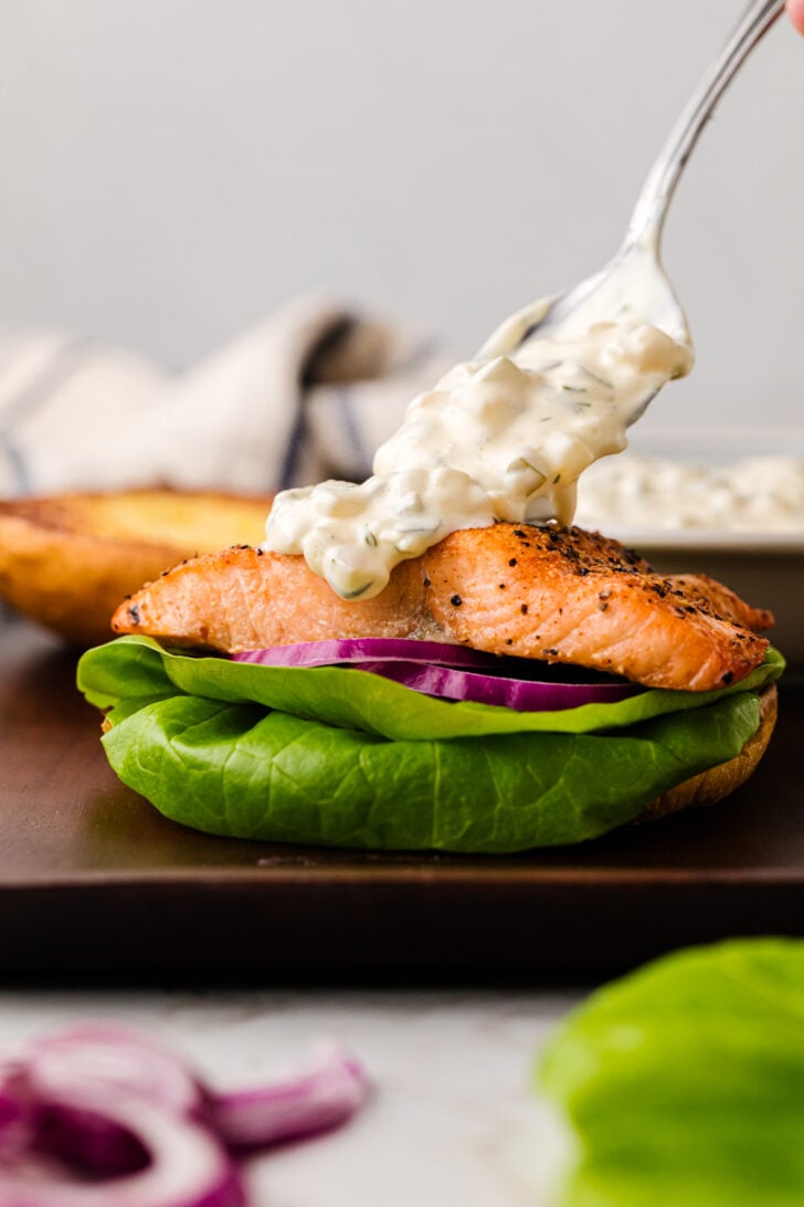 A spoon scooping creamy sauce onto a salmon sandwich. The sandwich has a piece of grilled salmon, bright green lettuce, and red onion slices on a bun. A halved bun and a piece of cloth are in the blurred background.