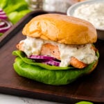 Salmon Sandwich: Fresh And Easy served on a toasted bun with creamy sauce, crispy salmon, lettuce and red onion on a wooden board