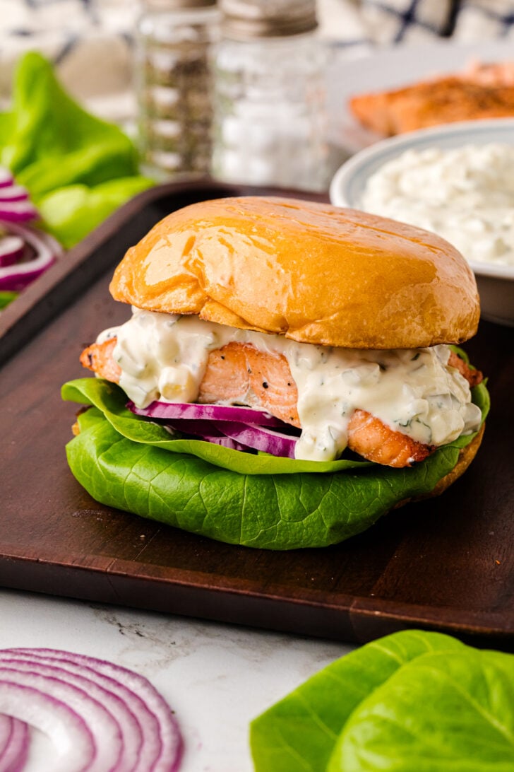 Close-up of a sandwich with salmon, creamy white sauce, lettuce, and red onion on a wooden board. The sandwich is made with a shiny brioche bun.