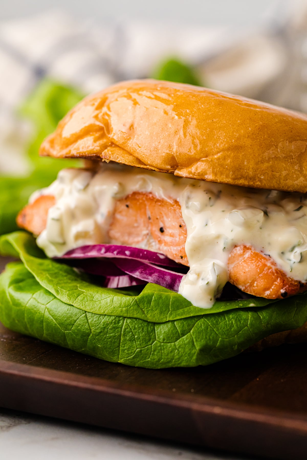 Salmon Sandwich: Fresh And Easy served on toasted bun with creamy sauce, red onion, and green lettuce on dark wooden board