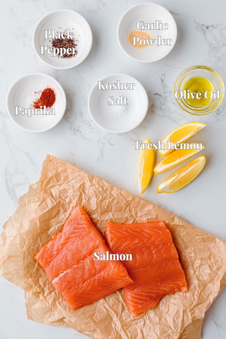 Top-down view of raw salmon on butcher paper with seasonings in small bowls: black pepper, paprika, kosher salt, garlic powder, and olive oil. Fresh lemon wedges are next to the salmon on a light marble surface.