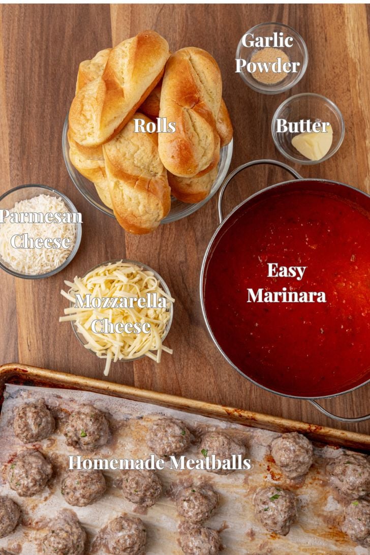 An overhead view of ingredients for meatball subs, including rolls, garlic powder, butter, parmesan cheese, mozzarella cheese, a pot of marinara sauce labeled "Easy Marinara," and a baking sheet with homemade meatballs.