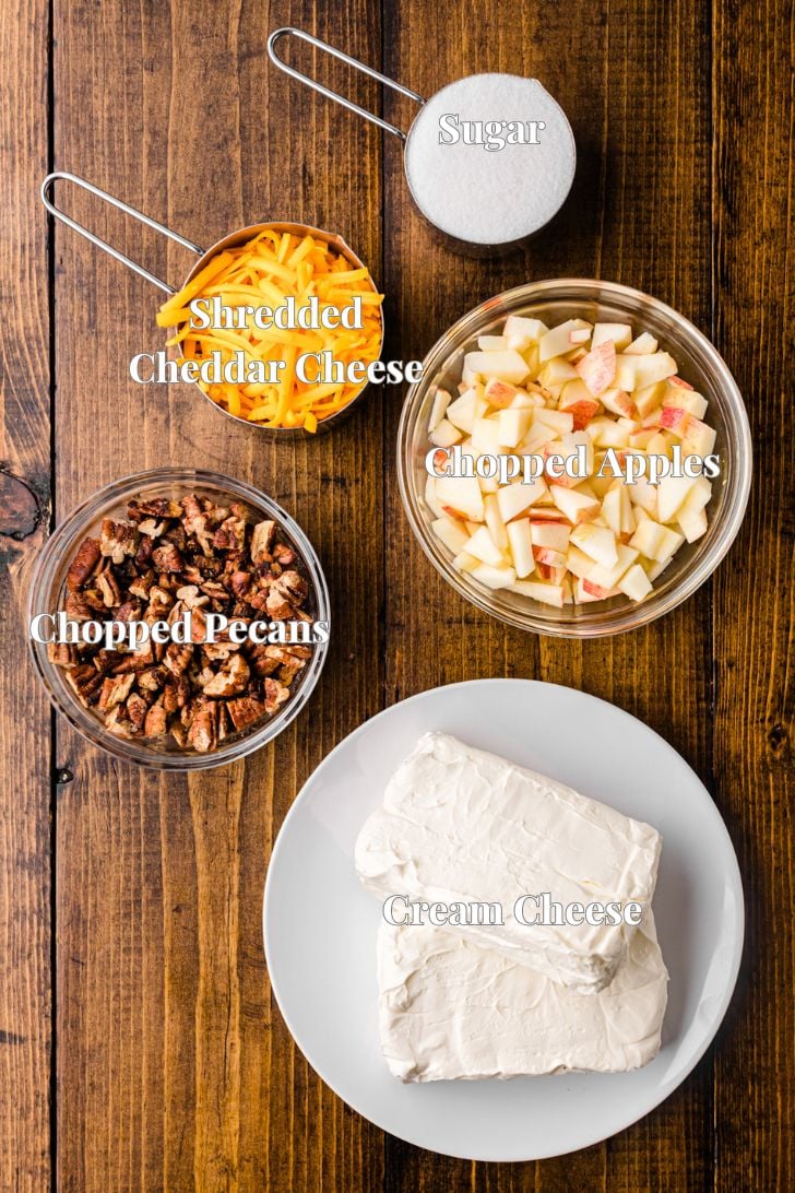 Top view of a wooden table with five ingredients in bowls: shredded cheddar cheese, chopped apples, chopped pecans, sugar, and cream cheese on a plate, all labeled.