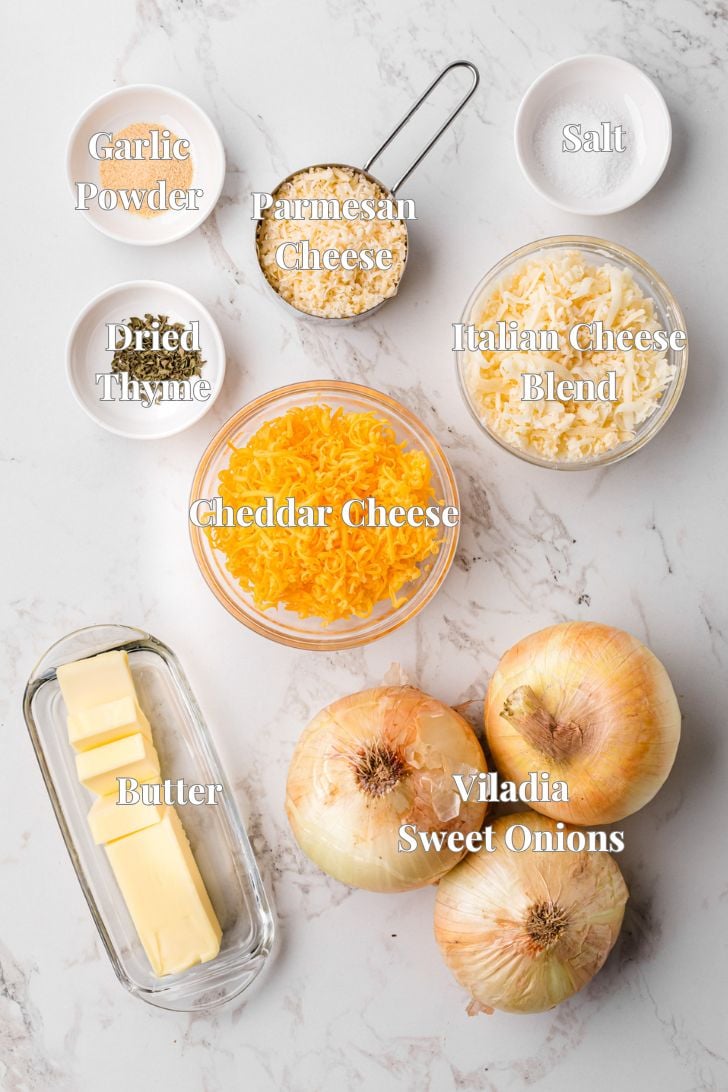 Ingredients for a recipe are laid out on a marble surface. Items include garlic powder, dried thyme, butter, salt, Parmesan cheese, Italian cheese blend, cheddar cheese, and Vidalia sweet onions. Each is labeled with text.