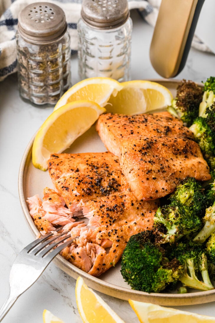 A plate with seasoned salmon fillets and roasted broccoli. Lemon wedges are placed around the plate. In the background, there are salt and pepper shakers and a striped cloth. A fork is flaking a piece of salmon.