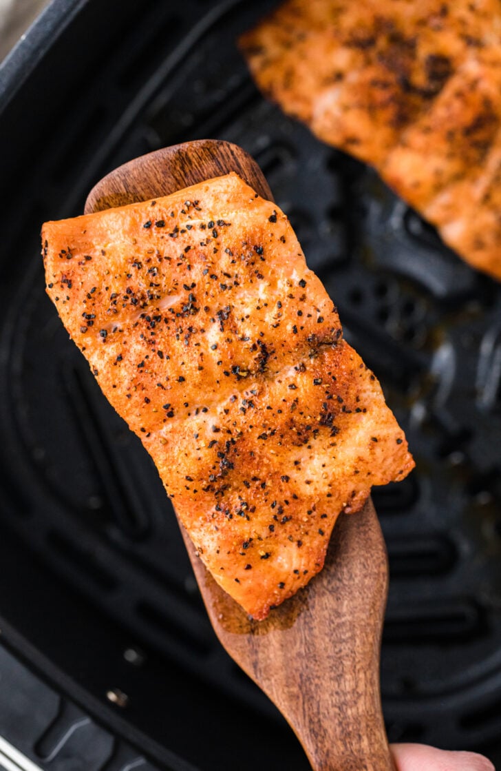 A piece of seasoned, cooked fish on a wooden spatula, held over a black grill pan. The fish features a golden-brown, crispy surface with visible seasoning. Another piece of fish is partially visible in the background.