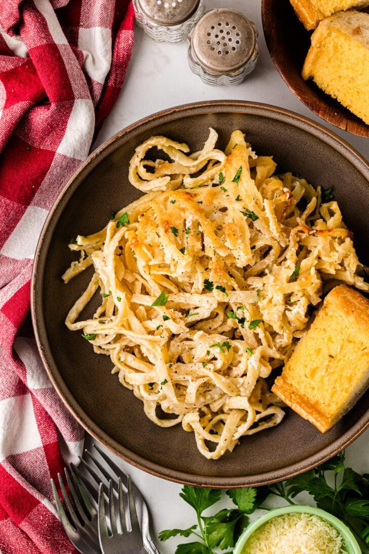 A glass baking dish filled with creamy baked spaghetti topped with melted cheese and chopped parsley. A decorative silver fork is partially inserted into the spaghetti, and a red-and-white cloth is partially visible on the side.