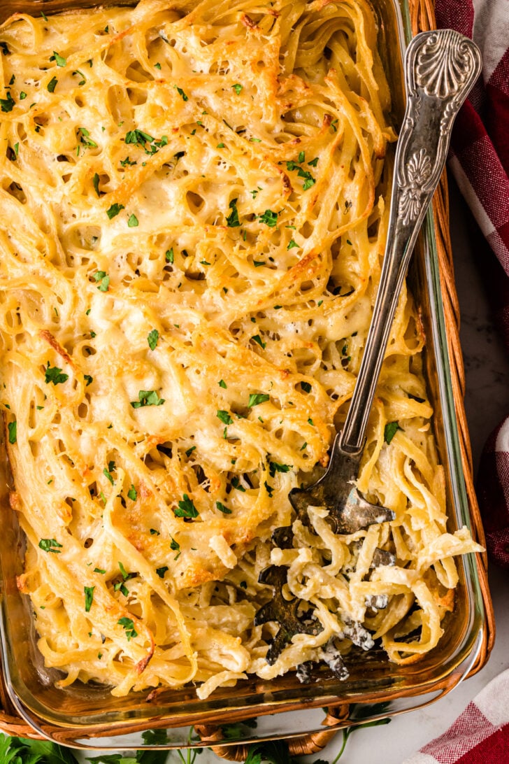 A glass baking dish filled with creamy baked spaghetti topped with melted cheese and chopped parsley. A decorative silver fork is partially inserted into the spaghetti, and a red-and-white cloth is partially visible on the side.