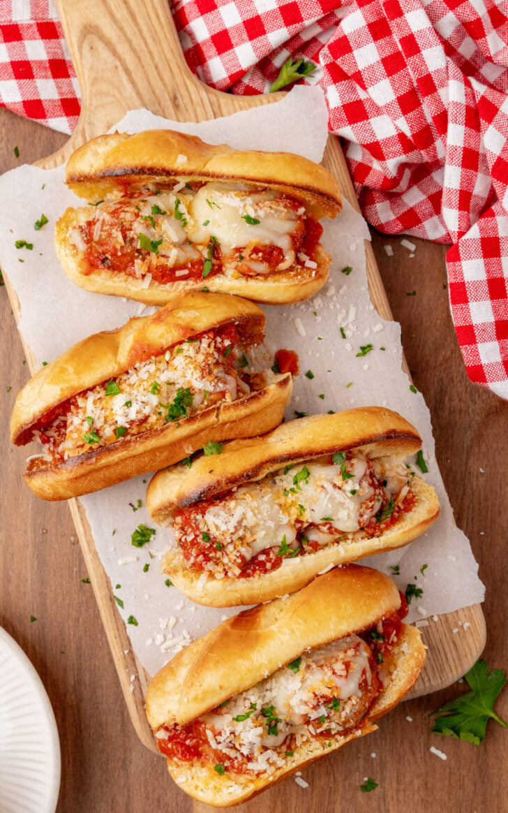Four meatball subs with melted cheese and marinara sauce on a wooden board lined with parchment paper. The subs are garnished with chopped parsley and grated cheese. A red and white checkered cloth is partially visible beside the board.