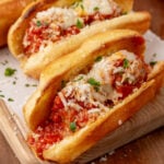 Meatball subs topped with melted cheese, red sauce, and fresh parsley on a wooden board with red checkered napkin