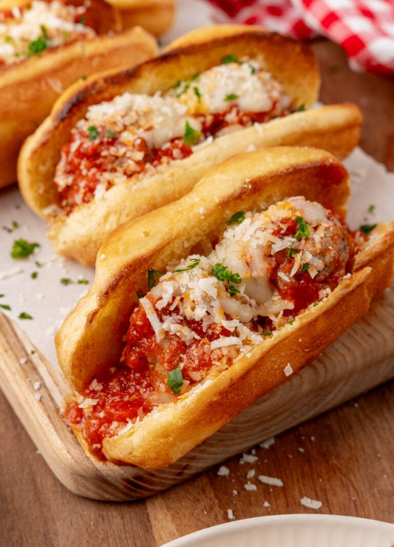 Homemade meatball subs 5 on wooden board topped with melted cheese, marinara sauce, and fresh parsley garnish