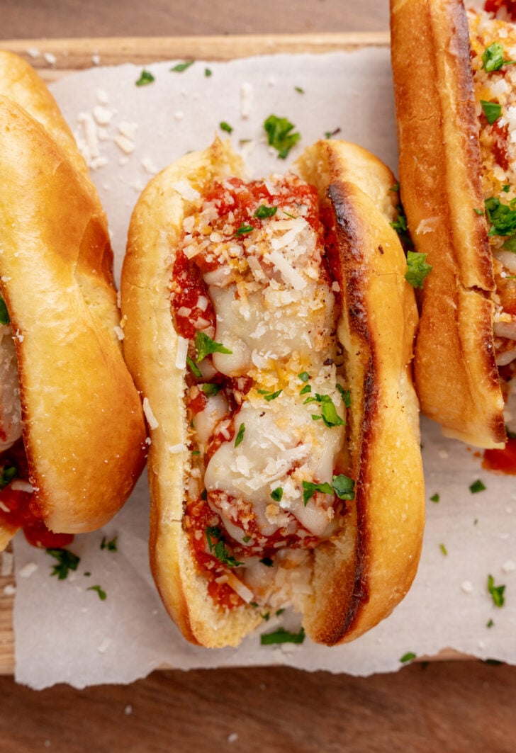 Two toasted sandwiches filled with melted cheese, tomato sauce, and garnished with grated Parmesan and chopped parsley on white parchment paper.