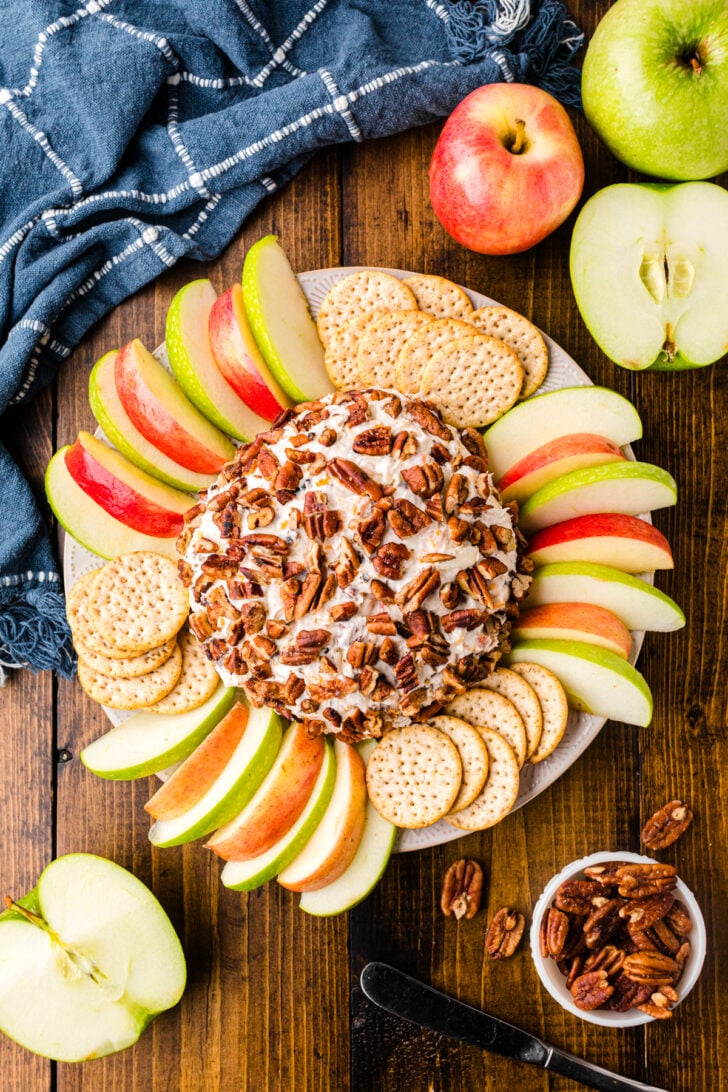 A cheese ball coated with pecans sits on a plate surrounded by apple slices and round crackers. A blue cloth is in the corner, with whole apples and a small bowl of pecans nearby on a wooden surface.