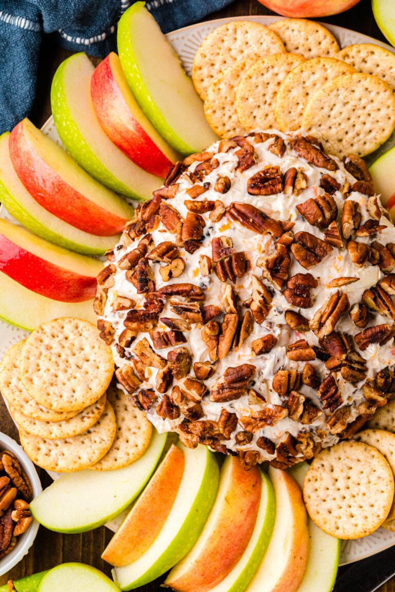 Party Cheese Ball with pecans on white plate surrounded by apple slices and crackers, overhead view