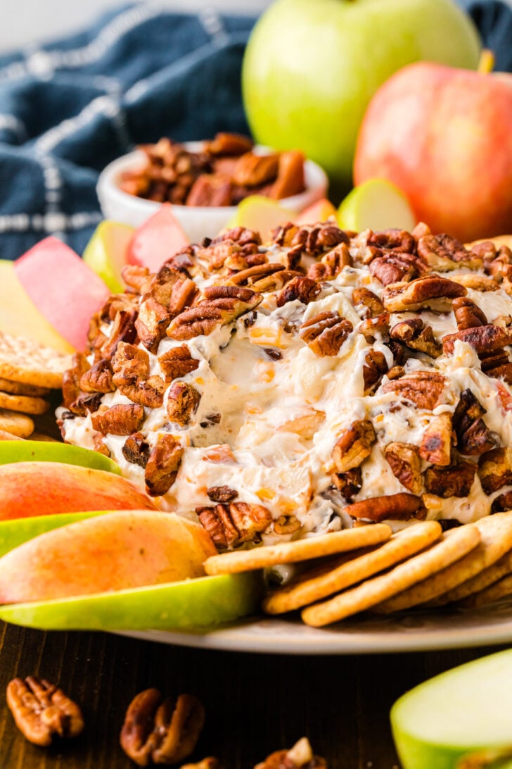 A cheese ball covered with pecans is surrounded by apple slices and crackers on a plate. In the background, there are whole apples, pecans, and a blue checkered cloth.