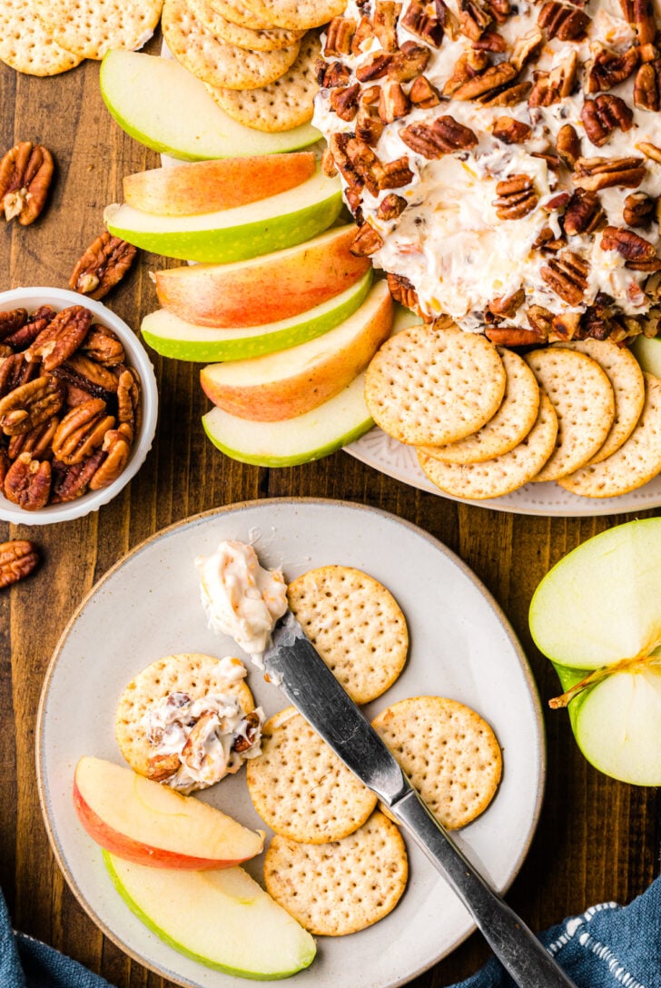 A plate of round crackers topped with a creamy pecan mixture, next to apple slices. A cheese ball covered with pecans, additional crackers, and apple slices are on a wooden table. A small bowl of pecans is nearby.