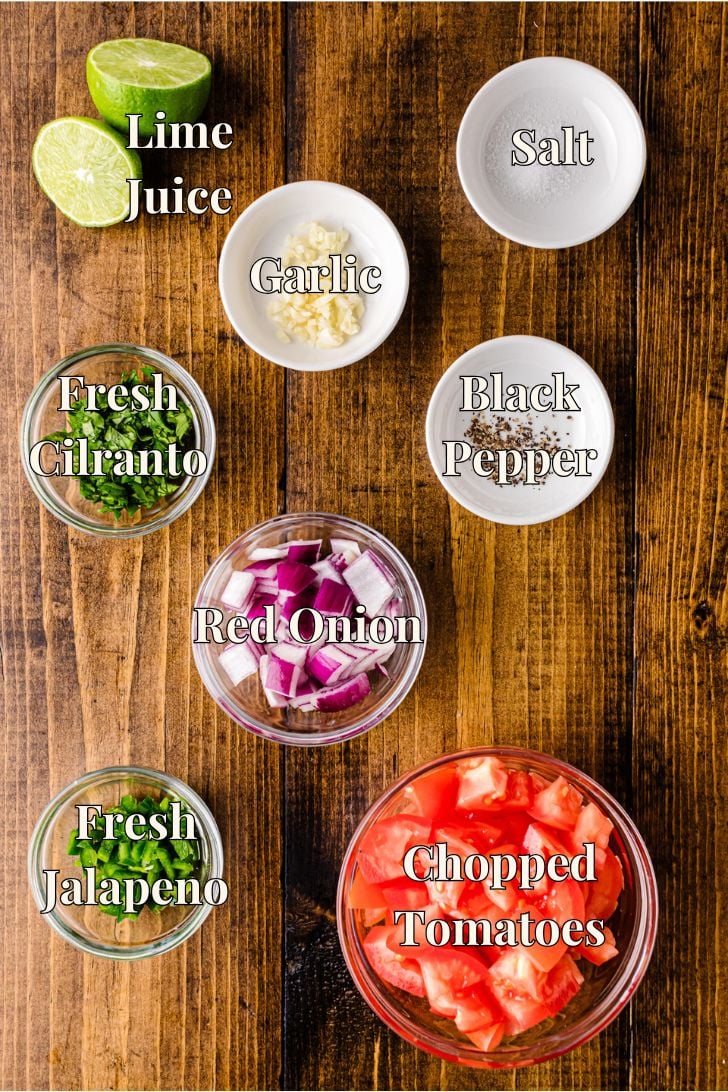 Overhead view of ingredients for salsa on a wooden surface: a halved lime, sliced garlic, a bowl of salt, chopped cilantro, black pepper, diced red onion, chopped jalapeños, and diced tomatoes, each labeled.