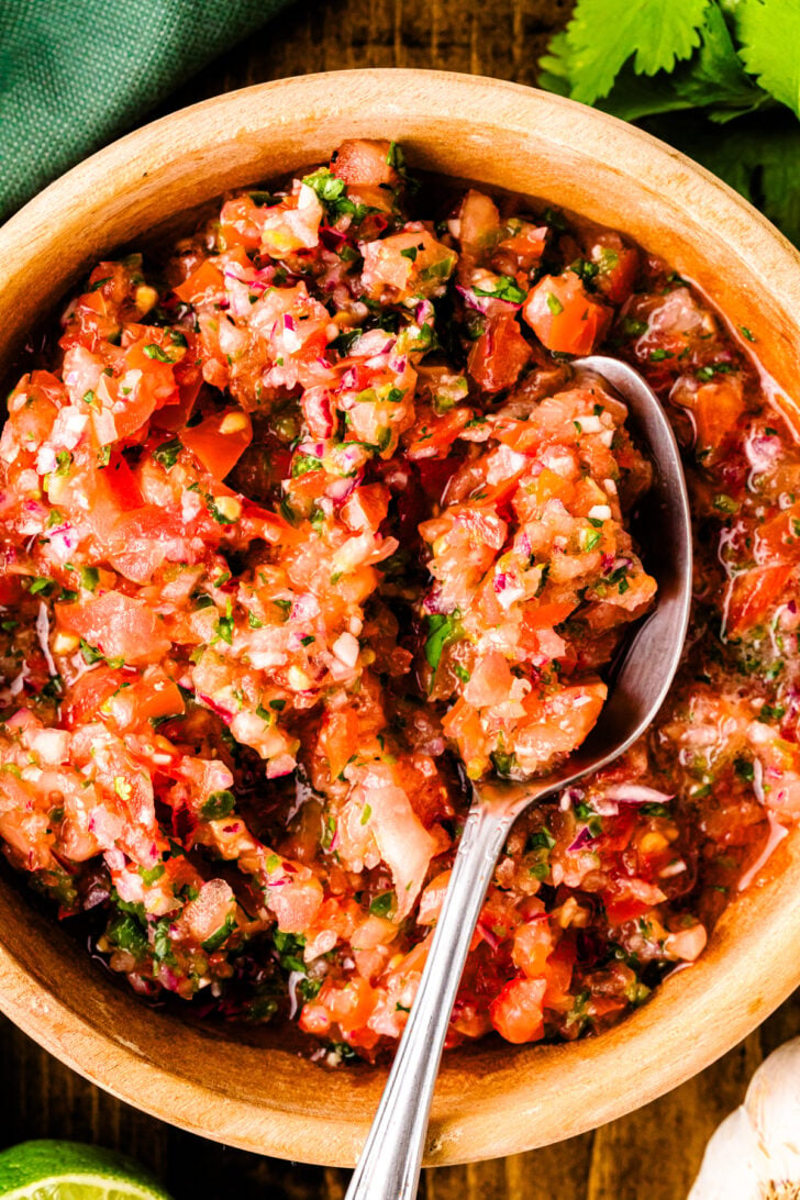 A close-up of a wooden bowl filled with chunky salsa made from chopped tomatoes, onions, cilantro, and other fresh ingredients, with a spoon resting inside.
