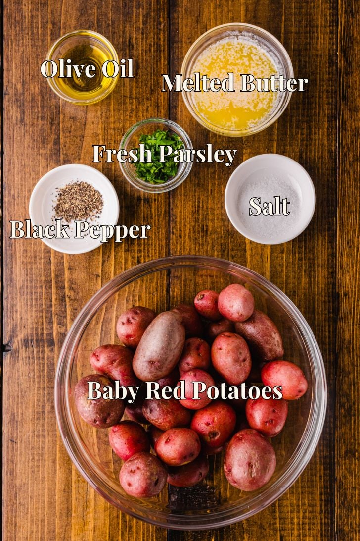 A wooden table with ingredients in bowls: baby red potatoes, olive oil, melted butter, fresh parsley, black pepper, and salt. Labels indicate each ingredient.