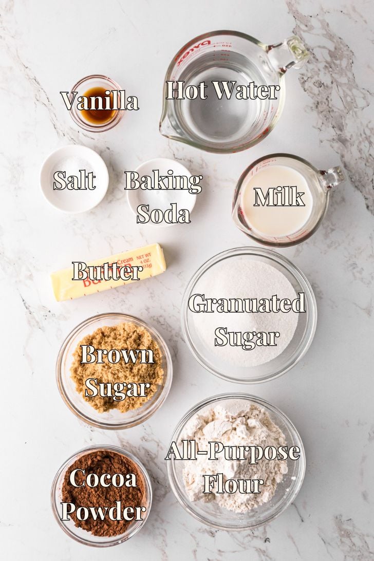 Top-down view of labeled baking ingredients on a marble surface: vanilla, hot water, milk, salt, baking soda, butter, granulated sugar, brown sugar, cocoa powder, and all-purpose flour in glass bowls and measuring cups.