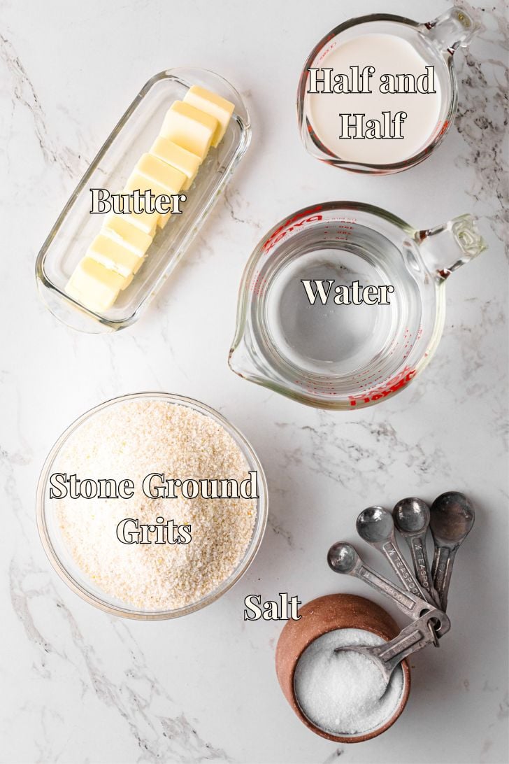 A top view of labeled ingredients for grits on a marble surface: half and half in a measuring cup, water in a measuring cup, sliced butter, a bowl of stone ground grits, a small bowl of salt, and measuring spoons.