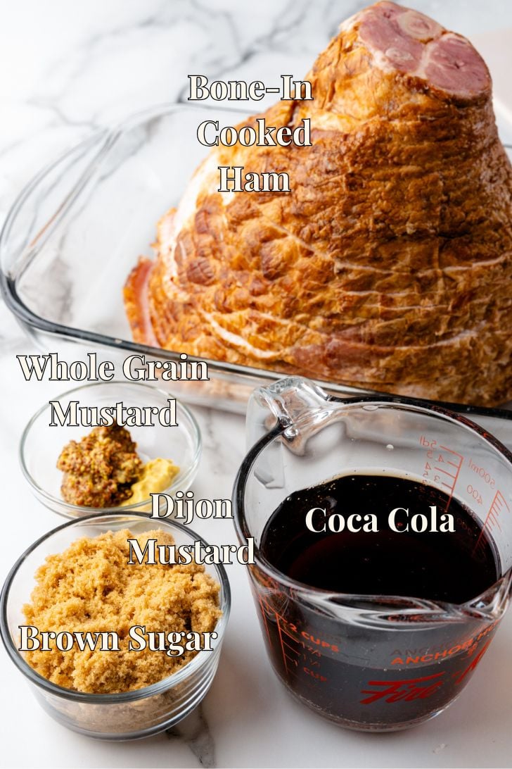 A bone-in cooked ham in a glass dish is surrounded by small bowls of whole grain mustard, Dijon mustard, and brown sugar. A measuring cup filled with Coca-Cola completes the glaze ingredients in the image.