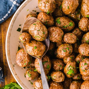 Roasted af baby potatoes 5 garnished with fresh parsley in cream bowl with gold Greek key pattern, served with spoon on dark wood table