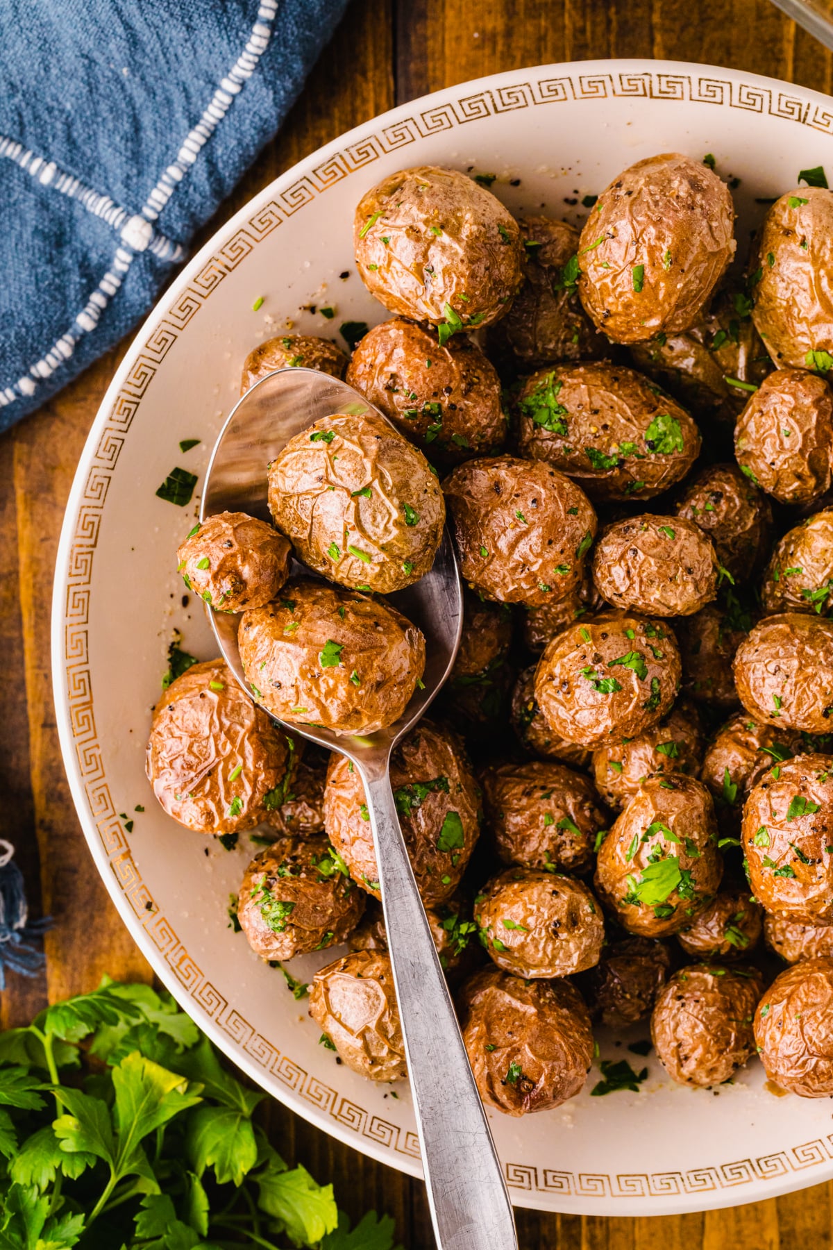 Roasted af baby potatoes 5 garnished with fresh parsley in cream bowl with gold Greek key pattern, served with spoon on dark wood table