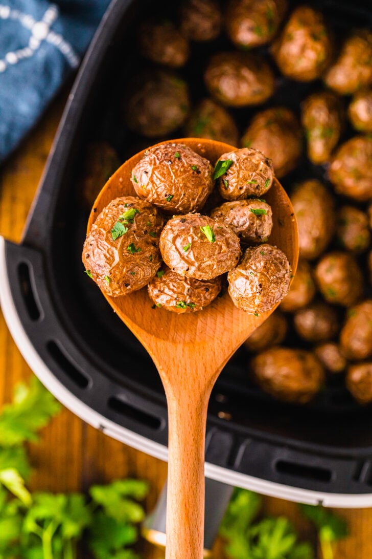 Wooden spoon holding roasted af baby potatoes 9 garnished with fresh herbs over cast iron skillet with more potatoes