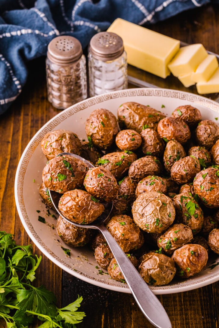 A bowl of roasted baby potatoes garnished with chopped parsley and a spoon. In the background, butter sticks, salt and pepper shakers, and a blue cloth are visible. Fresh parsley is on the wooden table.